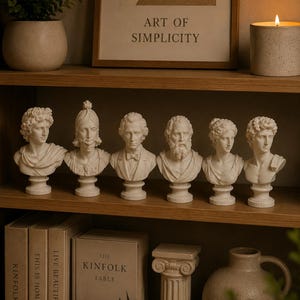 Puede incluir: Exhibición de bustos blancos de figuras históricas sobre una estantería de madera, junto a libros, una columna decorativa y un jarrón. Un cuadro enmarcado con el texto "ART OF SIMPLICITY" y una vela encendida.