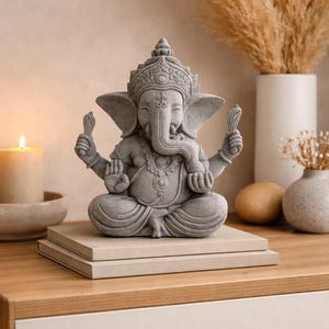 Op de afbeelding: Een grijze stenen beeld van Ganesha, de hindoeïstische godheid, zittend in een meditatieve houding. De olifantenkopfiguur is versierd met ingewikkelde details en sieraden. Het beeld staat op een stapel boeken op een houten tafel.