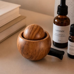 May include: A wooden car air freshener with a spherical top and a bowl-shaped base. The air freshener is brown and has a clip for attachment. Two amber glass bottles of essential oil are in the background.