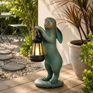 Può includere: Statua di coniglio turchese che tiene in mano una lanterna nera con luce gialla calda. La statua ha un aspetto invecchiato e poggia su una base di pietra. Decorazione da giardino all'aperto circondata dal verde.