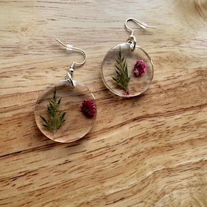 May include: Clear resin earrings with silver hooks. Each earring features a small sprig of green foliage and a small, dried red flower. The earrings are round and hang from silver-colored ear wires. The background is a wooden surface.