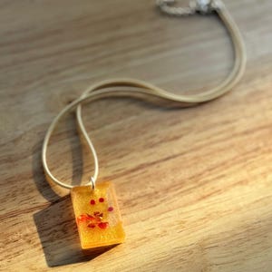 Puede incluir: Un collar con colgante rectangular con base de resina amarillo claro, incrustado con elementos florales rojos y naranjas. El colgante está suspendido de un collar de cordón color crema. El collar descansa sobre una superficie de madera.