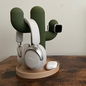 Op de afbeelding: Een groene cactusvormige standaard houdt een witte koptelefoon, een smartwatch en een witte draadloze oortelefooncase vast. De standaard heeft een beige basis en is gemaakt van een textuurrijk materiaal. De koptelefoon is voorzien van de naam "BOSE".
