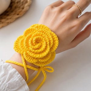 May include: A bright yellow, crocheted flower wrist cuff. The cuff is designed to look like a rose, with multiple layers of petals. The cuff is tied with yellow yarn. The cuff is worn on a wrist.