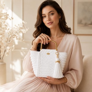 May include: A white crocheted handbag featuring a structured trapezoidal shape, a gold-toned metal top handle, and a matching white tassel charm attached to a gold ring. The bag has a textured knit pattern and a small gold-toned oval brand plaque.