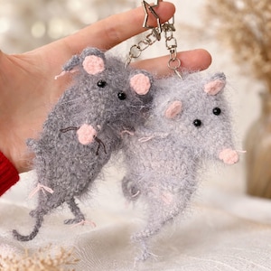 May include: Two grey, fluffy mouse keyrings with pink ears and noses. The keyrings are attached to a silver metal chain and star-shaped clasp. The mice have black bead eyes and small whiskers.