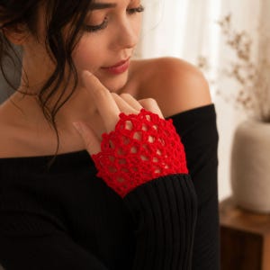 May include: A bright red fingerless glove featuring an intricate, open-work crochet pattern with a scalloped edge. The glove covers the back of the hand and extends into a ribbed black sleeve, showcasing a detailed, textured yarn construction.