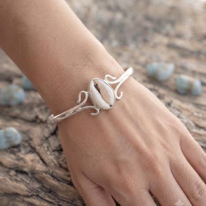 Op de afbeelding: Een zilveren armband met een witte kaurischelp in het midden. De metalen band heeft een gepolijste afwerking en decoratieve krulvormige metalen details aan weerszijden van de schelp, wat zorgt voor een open ontwerp rond de pols.