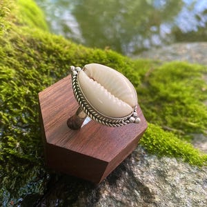 Puede incluir: Un anillo que presenta una concha de cauri de color crema lisa engastada en un marco de plata pulida. El engaste está decorado con un borde trenzado similar a una cuerda y pequeñas cuentas de plata en los extremos, creando un diseño de banda metálica detallado.