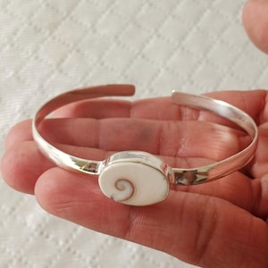 Op de afbeelding: Een zilveren manchetarmband met een gepolijste, ovaalvormige witte schelp in het midden. De schelp vertoont een natuurlijk, bruin spiraalpatroon. De metalen band is glad, reflecterend en heeft een open uiteinde voor om de pols.