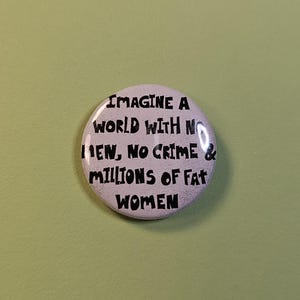 Puede incluir: Un botón blanco con texto negro que dice "IMAGINE A WORLD WITH NO MEN, NO CRIME & MILLIONS OF FAT WOMEN". El botón es redondo y está sobre una superficie verde claro.
