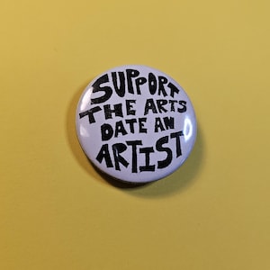May include: A white button with black text that reads "SUPPORT THE ARTS DATE AN ARTIST." The button is round and set against a yellow background. The text is in a bold, handwritten style.