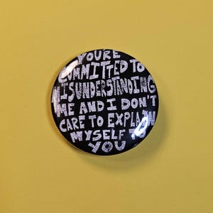 Peut inclure: Un bouton rond noir avec du texte blanc. Le texte dit : "YOU'RE COMMITTED TO MISUNDERSTANDING ME AND I DON'T CARE TO EXPLAIN MYSELF TO YOU." Le bouton est posé sur un fond jaune.