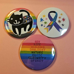 May include: Three circular badges with vibrant designs. One displays a black cat on a rainbow backdrop with the text "Pride is a Protest." Another shows a blue ribbon with the text "Protect Queer Youth" and stars. The third has rainbow stripes and the words "Sex, Life, Healing, Sunlight, Nature, Magic, Serenity, Spirit."
