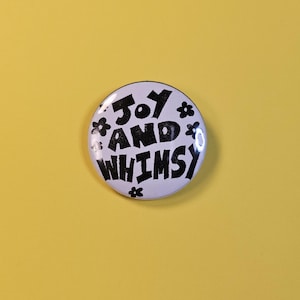 Pode incluir: Um botão branco com as palavras "JOY AND WHIMSY" em preto, rodeado por pequenas ilustrações de flores. O botão está sobre um fundo amarelo liso, criando uma estética alegre e divertida, ideal para acessórios.