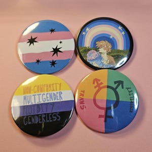 Op de afbeelding: Vier ronde buttons met kleurrijke ontwerpen. Eén toont de transgender vlag met sterren, een andere een cartoon karakter met een regenboog, en een derde tekst over genderidentiteit. De laatste button heeft het trans symbool.