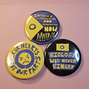 Peut inclure: Trois badges ronds avec du texte et des graphiques. L'un porte l'inscription "INTERSEX REPRESENTATION AND RECOGNITION NOW" avec un design jaune et bleu. Un autre dit "FIX UR HEARTS NOT OUR PARTS" avec un visage triste. Le troisième dit "BIOLOGY WAS NEVER BINARY".