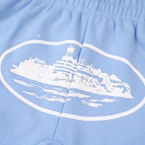 K&ouml;nnte beinhalten: Hellblaue Shorts mit einem wei&szlig;en ovalen Aufdruck, der einen Leuchtturm auf einer felsigen Insel zeigt. Die Shorts haben einen elastischen Bund und sind aus einem weichen, bequemen Stoff gefertigt.