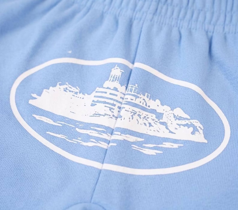 K&ouml;nnte beinhalten: Hellblaue Shorts mit einem wei&szlig;en ovalen Aufdruck, der einen Leuchtturm auf einer felsigen Insel zeigt. Die Shorts haben einen elastischen Bund und sind aus einem weichen, bequemen Stoff gefertigt.