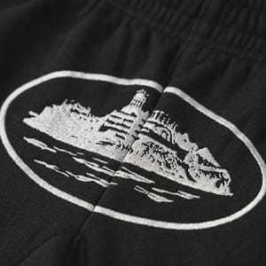 May include: Black sweatpants with a white embroidered design of a building on a rocky island, surrounded by water, inside an oval frame. The design is detailed and textured.