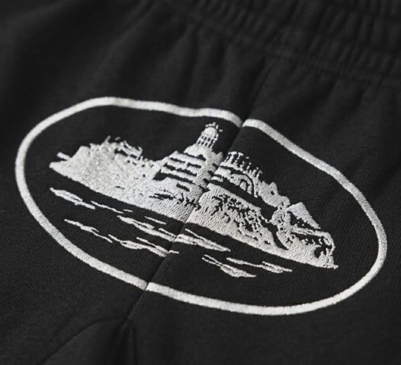 May include: Black sweatpants with a white embroidered design of a building on a rocky island, surrounded by water, inside an oval frame. The design is detailed and textured.