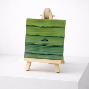 May include: A small artwork on a wooden easel. The artwork depicts a green landscape with a blue car. The artist's name, WITHER INOMONORST, is printed at the bottom of the artwork.