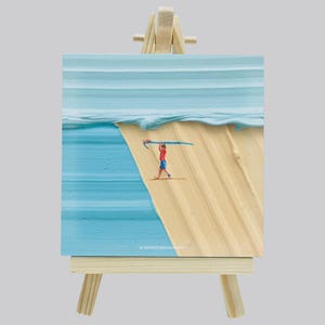 May include: A painting on canvas depicting a beach scene with a surfer carrying a surfboard. The artwork is displayed on a small wooden easel. The painting features a blue ocean, sandy beach, and a figure in red and blue. The artist's signature is visible.