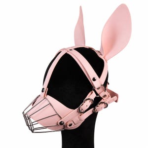May include: A light pink head harness featuring two tall, upright rabbit ears attached to the top. The headgear includes a series of adjustable straps with silver-toned metal buckles and a wire cage muzzle covering the front nose area.