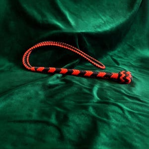 Puede incluir: Un látigo de cuero rojo y negro con un mango trenzado y un extremo anudado. El látigo está curvado y descansa sobre una superficie de terciopelo verde oscuro. El diseño del látigo presenta secciones rojas y negras alternas.