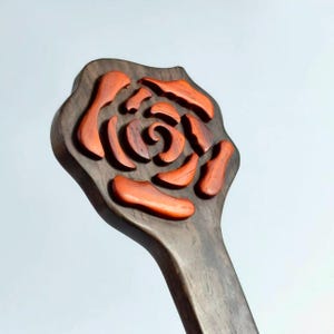 May include: A wooden carving of a rose. The rose is made of reddish-orange pieces set into a dark brown, paddle-shaped base. The carving is detailed, with the rose petals and center clearly defined. The handle is also made of dark brown wood.