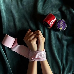 Peut inclure: Ruban adhésif rose autour des poignets, avec un rouleau de ruban rose et deux rouleaux de ruban rouge et noir. Le rouleau de ruban noir contient du texte et un graphique. Le fond est un tissu de velours vert foncé.