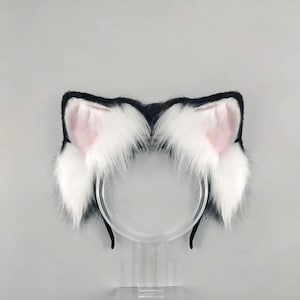 Puede incluir: Una diadema con orejas de gato. Las orejas son negras con pelaje blanco y suave y el interior rosa. La diadema es de plástico transparente, sobre un fondo gris claro.