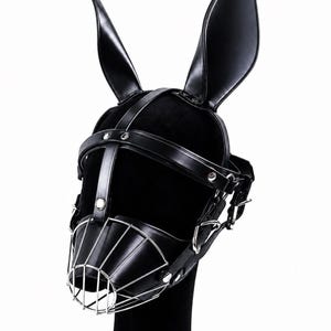 Peut inclure: Un accessoire de tête noir doté de grandes oreilles de lapin droites en matériau lisse aspect cuir. Le masque comprend des sangles réglables avec des rivets en métal argenté et une muselière en cage faite de fines barres métalliques polies.