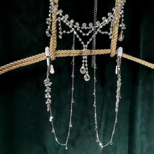 Puede incluir: Un conjunto de joyería corporal plateada con detalles de cristal colgantes. El conjunto incluye una pieza tipo collar y dos cadenas colgantes con colgantes de cristal. La joyería se exhibe sobre un fondo verde oscuro.