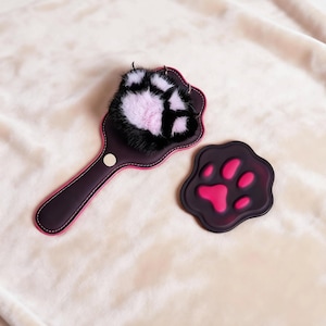 May include: A dark purple cat paw-shaped paddle with a pink trim and a fluffy black and pink paw print. A matching dark purple coaster with a pink paw print is also present. The items are on a soft, cream-colored surface.