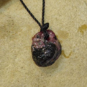 May include: A raw, irregularly shaped pendant featuring a deep red and black stone suspended from a thin black cord. The stone has a textured, unpolished surface with visible dark inclusions and a knot securing the cord at the top centre.