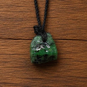 May include: A raw, uncut green gemstone pendant suspended from a black cord. The stone features a deep emerald hue with dark, irregular inclusions and a polished, glossy surface texture. The cord is knotted securely through the top of the stone.