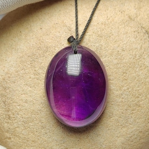 Op de afbeelding: Een gepolijste, ovale amethist hanger, hangend aan een zwart koord. De edelsteen vertoont een gradiënt van paarse tinten, van licht aan de basis tot diep violet aan de bovenkant. Een zilverkleurige metalen sluiting bevestigt het koord aan de hanger.