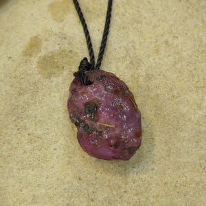 Op de afbeelding: Een ruwe, diep magenta gekleurde edelsteen hanger met een onregelmatig, getextureerd oppervlak en donkere insluitingen, hangend aan een dun, zwart gedraaid koord. De steen heeft een natuurlijke, ongepolijste uitstraling met zichtbare putjes.
