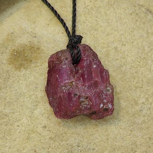 Peut inclure: Un pendentif en pierre brute de couleur magenta profond avec une texture cristalline rugueuse et des bords irréguliers. La pierre est suspendue à un fin cordon torsadé noir qui passe à travers un petit trou percé près du centre supérieur.