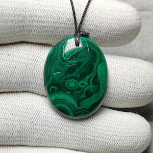 Puede incluir: Colgante ovalado de malaquita con un patrón verde arremolinado, suspendido de un cordón negro. La piedra pulida muestra bandas de verde claro y oscuro, creando un diseño natural y llamativo. Una pieza de joyería única.