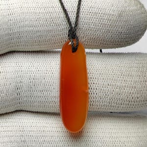 Op de afbeelding: Een gladde, langwerpige, doorschijnende oranje carneool hanger met een afgeronde onderkant. De steen heeft een klein gaatje aan de bovenkant, waar een dun, donker, gevlochten koord doorheen is geregen om een ketting te vormen. Het oppervlak is glanzend.