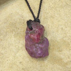 Peut inclure: Un pendentif en rubis brut non poli suspendu à un cordon noir torsadé. La pierre précieuse présente une teinte magenta et violette profonde avec une texture de surface rugueuse et irrégulière et des facettes cristallines naturelles.