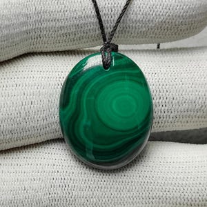 Puede incluir: Colgante ovalado de malaquita con un patrón verde arremolinado, suspendido de un cordón negro. La piedra pulida muestra un diseño natural vibrante. Una pieza de joyería única.