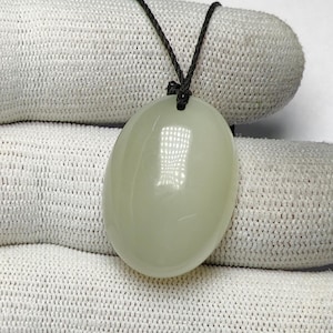 May include: An oval, pale green pendant necklace. The smooth, polished stone hangs from a dark cord. The pendant has a light, creamy colour and a glossy finish. The necklace is displayed against a white, textured background.