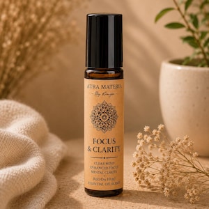 Może przedstawiać: Szklana butelka typu roll-on o pojemności 10 ml z czarną nakrętką i beżową etykietą. Na etykiecie znajduje się napis AURA MATERIA By Kinga, grafika mandali oraz słowa FOCUS & CLARITY, CLEAR MIND, ENHANCED FOCUS, MENTAL CLARITY i ESSENTIAL OIL BLEND.