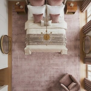 Puede incluir: Vista a&eacute;rea de un dormitorio con una gran alfombra rosa empolvado. La cama tiene ropa de cama blanca, almohadas rosa empolvado y una colcha decorativa. Dos mesitas de noche de madera flanquean la cama. Una l&aacute;mpara de ara&ntilde;a cuelga sobre la cama. Una silla a juego est&aacute; en la esquina.