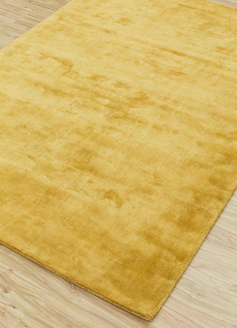 May include: A rectangular, golden-yellow rug with a soft, textured surface. The rug is made of a plush material and has a subtle sheen, suggesting a luxurious feel. The rug is placed on a light-colored wooden floor.