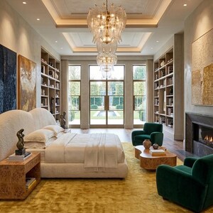 May include: A luxurious bedroom with a large bed, a yellow rug, and two green armchairs. A large chandelier hangs from the ceiling. Bookshelves and artwork adorn the walls. The room has a fireplace and large windows.
