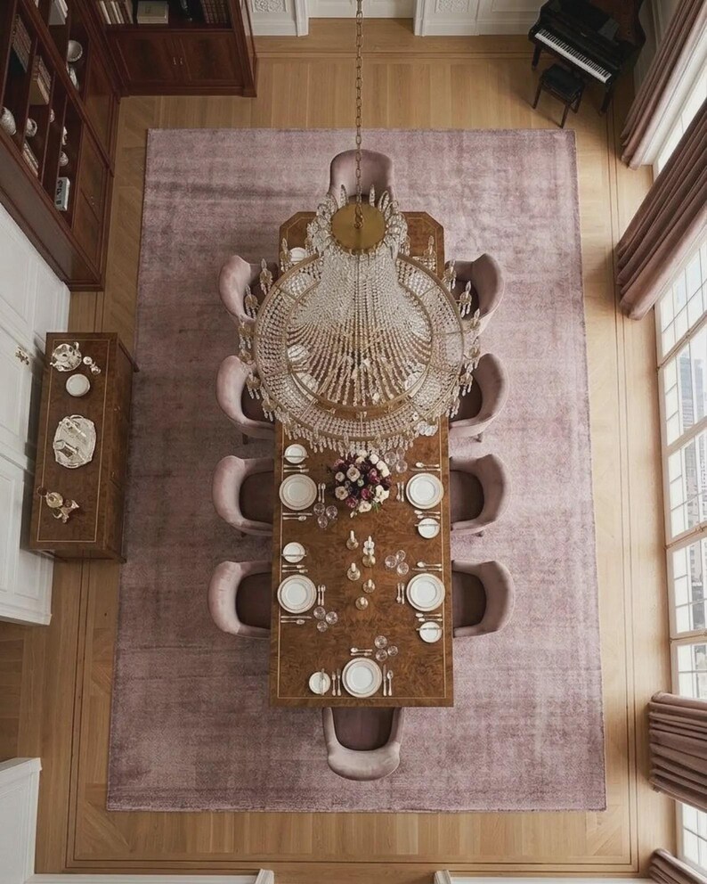 Puede incluir: Vista a&eacute;rea de un comedor formal. Una gran mesa rectangular de madera est&aacute; puesta con platos blancos y cristaler&iacute;a. Sillas rosas tapizadas rodean la mesa, que est&aacute; centrada en una gran alfombra malva. Una l&aacute;mpara de ara&ntilde;a de cristal cuelga encima.